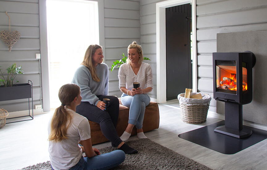 Tre tjejer som sitter på en brun sittpuff i ett grått vardagsrum framför braskaminen Jøtul F 167